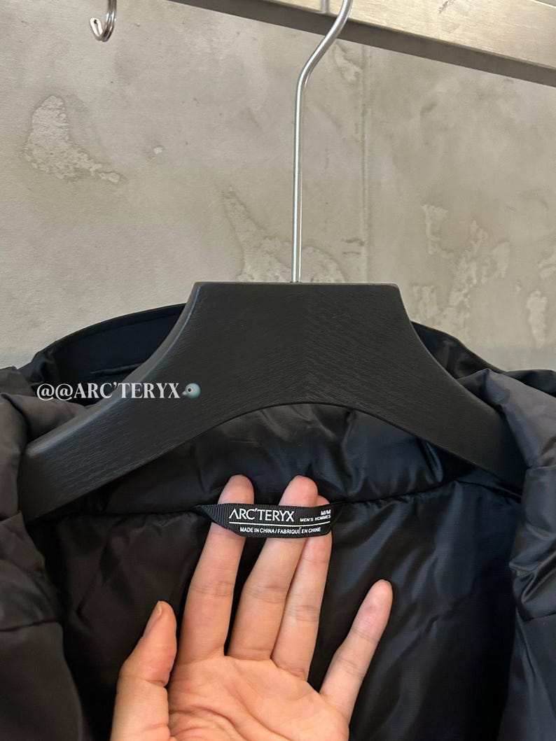 May include: Black jacket hanging on a black hanger. The jacket has a label that reads "ARC'TERYX" and "MADE IN CHINA". The jacket is made of a smooth, shiny material.