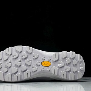 Puede incluir: Suela de zapatilla deportiva blanca con un patr&oacute;n texturizado y un logotipo "Vibram" amarillo. La suela est&aacute; dise&ntilde;ada para la tracci&oacute;n y la durabilidad, adecuada para actividades al aire libre. El calzado es probablemente para senderismo o trail running.