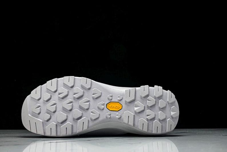 Puede incluir: Suela de zapatilla deportiva blanca con un patr&oacute;n texturizado y un logotipo "Vibram" amarillo. La suela est&aacute; dise&ntilde;ada para la tracci&oacute;n y la durabilidad, adecuada para actividades al aire libre. El calzado es probablemente para senderismo o trail running.