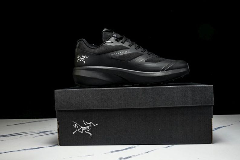 May include: A black athletic shoe, the Norvan 15, sits atop a black box. The shoe features a sleek design with the brand logo on the side. The box also has the brand logo in white. The background is black.