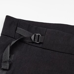 May include: Close-up of black pants with a black buckle and strap detail. The pants are made of a dark fabric and have a minimalist design. The focus is on the waistband and the functional strap.
