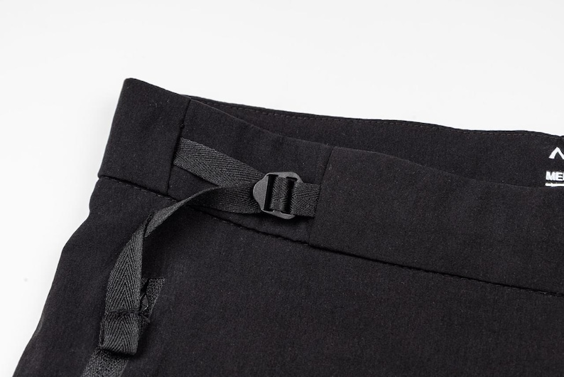 May include: Close-up of black pants with a black buckle and strap detail. The pants are made of a dark fabric and have a minimalist design. The focus is on the waistband and the functional strap.