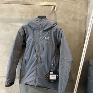 May include: A gray hooded jacket with a zipper closure and multiple pockets. The jacket has the brand logo on the chest and a tag with the GORE-TEX logo. The jacket is hanging on a metal bar.