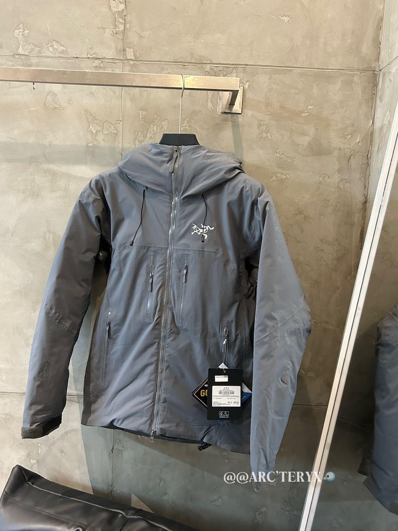 May include: A gray hooded jacket with a zipper closure and multiple pockets. The jacket has the brand logo on the chest and a tag with the GORE-TEX logo. The jacket is hanging on a metal bar.