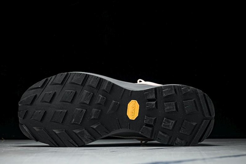 May include: Close-up of the sole of a beige and black athletic shoe. The black sole has a textured pattern with a yellow "Vibram" logo. The shoe is designed for outdoor activities.