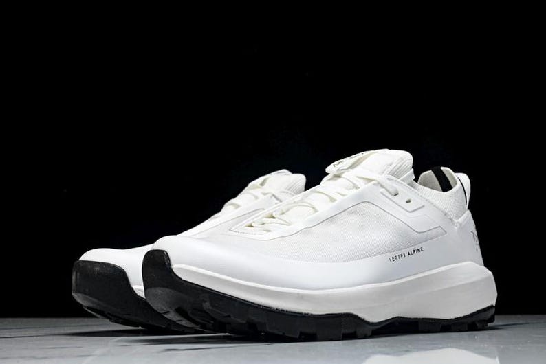 Puede incluir: Par de zapatillas deportivas blancas con suelas negras. Las zapatillas tienen un dise&ntilde;o aerodin&aacute;mico con el texto "VERTEX ALPINE" impreso en el lateral. Las zapatillas est&aacute;n sobre un fondo oscuro.