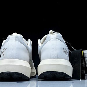 May include: Pair of white and light grey athletic shoes with a black sole and a cream-colored midsole. The shoes have a logo on the heel and a tag attached. The shoes are designed for outdoor activities.