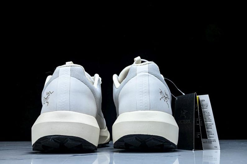 May include: Pair of white and light grey athletic shoes with a black sole and a cream-colored midsole. The shoes have a logo on the heel and a tag attached. The shoes are designed for outdoor activities.
