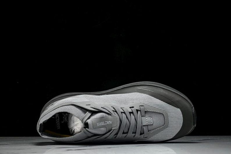 Puede incluir: Zapatilla deportiva gris con una parte superior de malla y una suela gris oscuro. El zapato tiene un cierre de cordones y la marca "Arc'teryx" es visible en la leng&uuml;eta. Dise&ntilde;ado para correr u otras actividades deportivas.