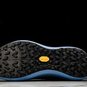 May include: Close-up of the sole of a running shoe. The sole is black with a textured pattern and a light blue midsole. A yellow "Vibram" logo is visible.