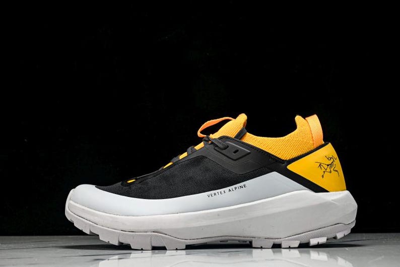 Puede incluir: Zapatilla deportiva negra y amarilla con suela blanca. La zapatilla tiene una parte superior negra con detalles amarillos en el tal&oacute;n y la leng&uuml;eta. Las palabras "VERTEX ALPINE" est&aacute;n impresas en el lateral.