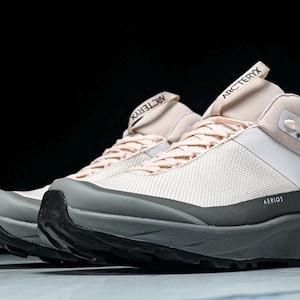 May include: A pair of light pink and white hiking shoes with gray and black accents. The shoes have the word "Arc'teryx" printed on the tongue and the word "Aerios" printed on the side. The shoes are designed for outdoor activities.