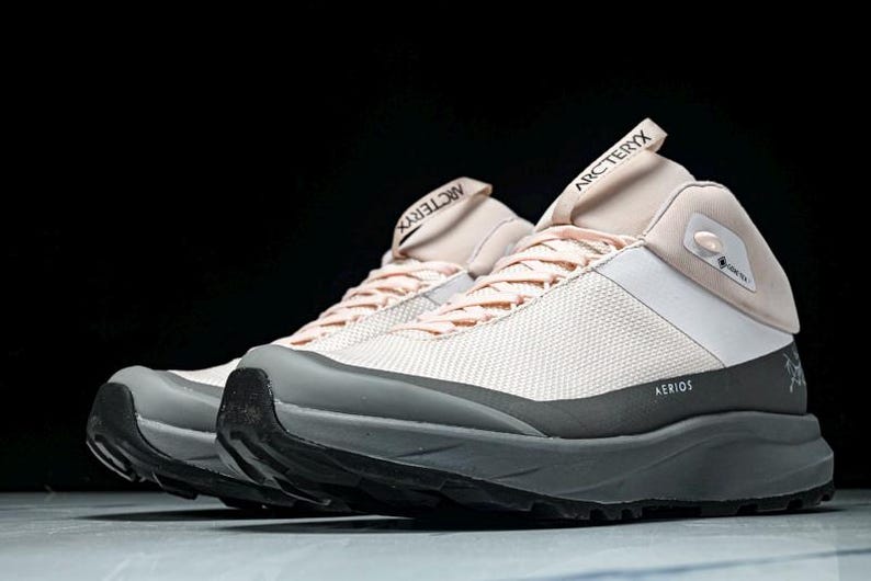 May include: A pair of light pink and white hiking shoes with gray and black accents. The shoes have the word "Arc'teryx" printed on the tongue and the word "Aerios" printed on the side. The shoes are designed for outdoor activities.