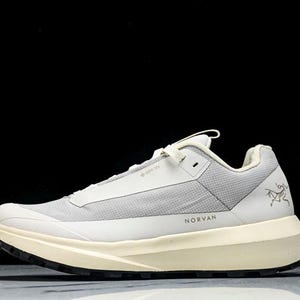 May include: A light gray and cream-colored athletic shoe with the word "NORVAN" printed on the side. The shoe has a mesh upper, a thick sole, and a black outsole. The Arc'teryx logo is visible on the side.