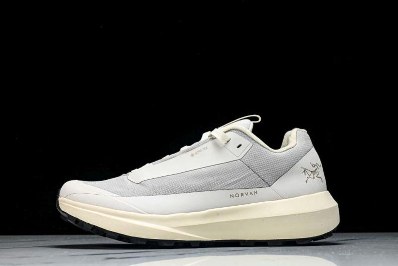 May include: A light gray and cream-colored athletic shoe with the word "NORVAN" printed on the side. The shoe has a mesh upper, a thick sole, and a black outsole. The Arc'teryx logo is visible on the side.