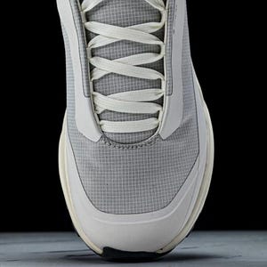 May include: Close-up of a gray and white athletic shoe. The shoe features a breathable mesh upper with white accents and laces. The sole is a combination of white and black. The design is modern and minimalist.