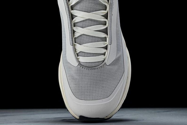 May include: Close-up of a gray and white athletic shoe. The shoe features a breathable mesh upper with white accents and laces. The sole is a combination of white and black. The design is modern and minimalist.