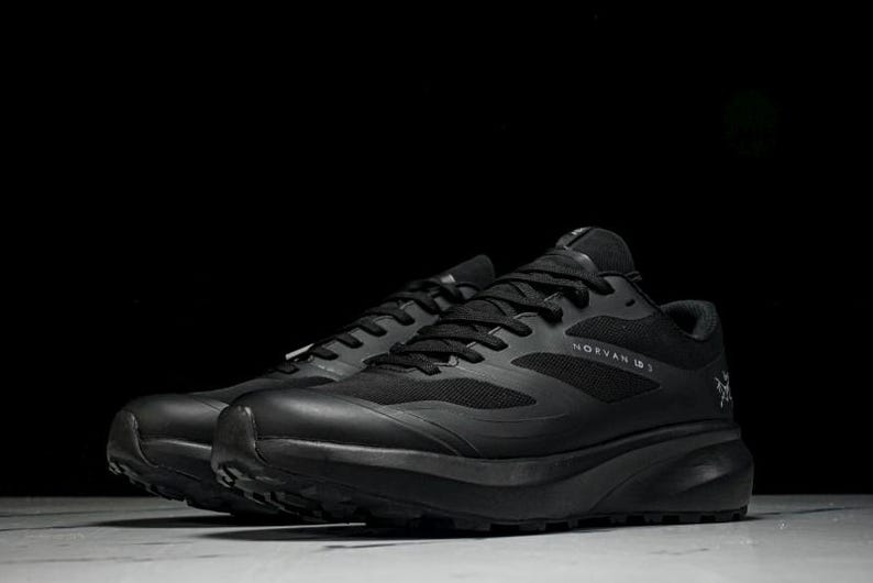 May include: Pair of black athletic shoes with the text "NORVAN IS" on the side. The shoes have a mesh upper, black laces, and a thick, textured sole. The shoes are designed for running or trail activities.