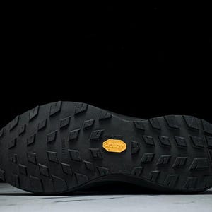 May include: Black athletic shoe sole with a textured pattern and a yellow Vibram logo. The sole features a diamond-shaped tread design for enhanced grip and durability. The shoe is designed for outdoor activities.