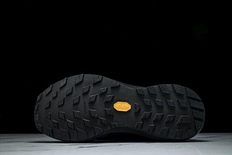 May include: Black athletic shoe sole with a textured pattern and a yellow Vibram logo. The sole features a diamond-shaped tread design for enhanced grip and durability. The shoe is designed for outdoor activities.