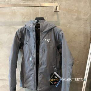 May include: A gray hooded jacket with a zipper closure and multiple pockets. The jacket has a white logo on the chest and a Gore-Tex tag. The jacket is hanging on a metal bar.