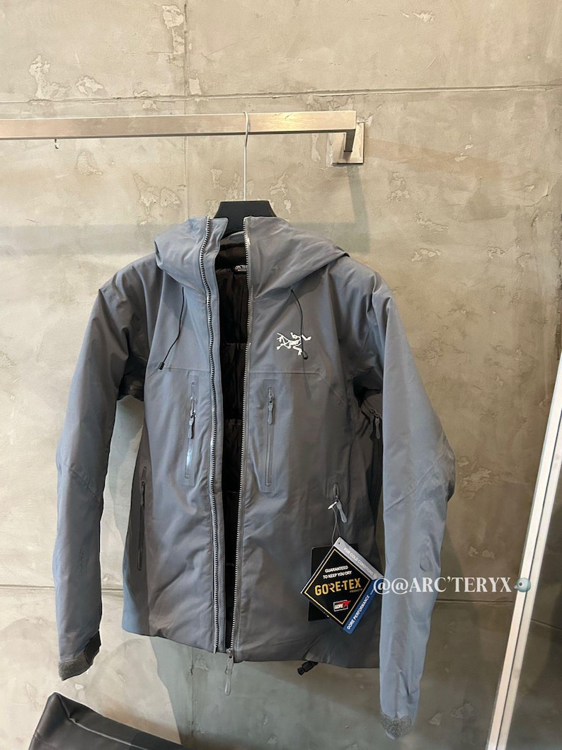 May include: A gray hooded jacket with a zipper closure and multiple pockets. The jacket has a white logo on the chest and a Gore-Tex tag. The jacket is hanging on a metal bar.