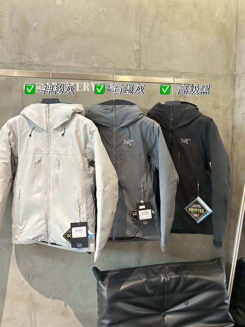 May include: Three hooded jackets in light gray, gray, and black hang on a rack. Each jacket has a zipper closure, pockets, and a brand logo on the chest. The jackets have tags attached. The text above the jackets is in Chinese.