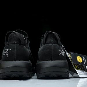 Puede incluir: Un par de zapatillas deportivas negras, vistas desde atr&aacute;s, con suela texturizada y un logotipo en el tal&oacute;n. Las zapatillas tienen una etiqueta con texto y un logotipo amarillo.