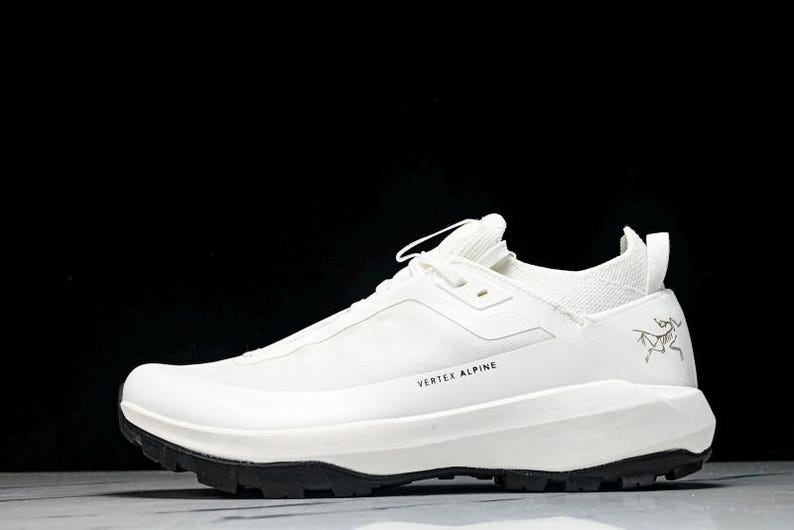 Puede incluir: Zapatilla deportiva blanca con suela negra. La zapatilla tiene el texto "VERTEX ALPINE" y un logotipo. Dise&ntilde;o aerodin&aacute;mico, probablemente para actividades al aire libre.