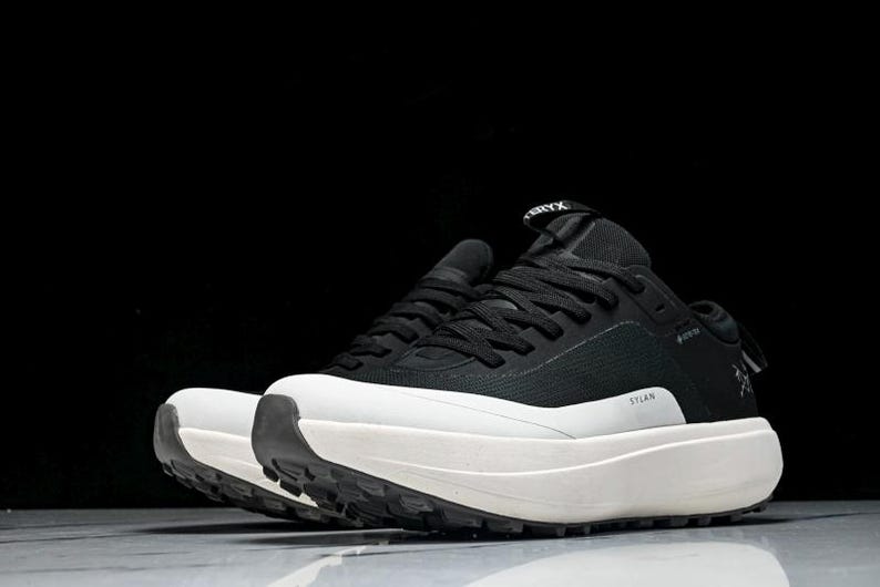 May include: Black and white athletic shoes with a thick, white sole. The shoes have black laces and a black upper with the word "STLAN" printed on the side. The brand name is visible on the tongue.