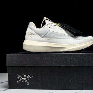 May include: A light gray and white athletic shoe with a black sole, sitting on a black box. The shoe has the word "NORVAN" printed on it. The box has a white logo on the front.