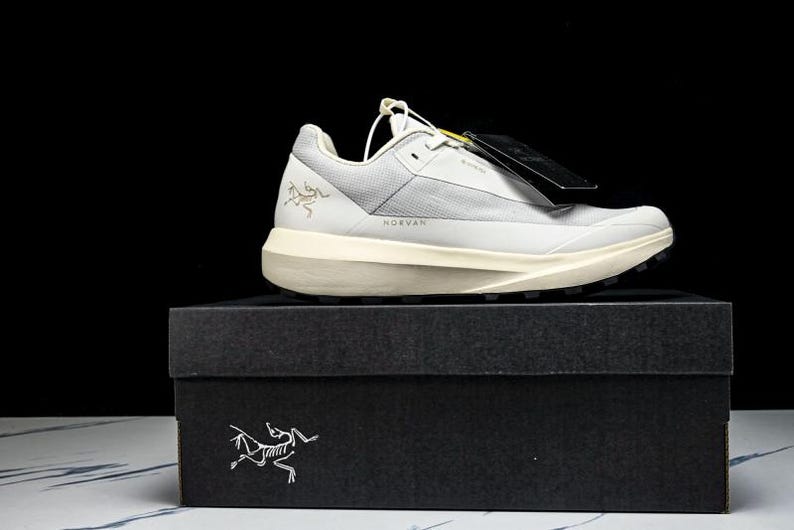 May include: A light gray and white athletic shoe with a black sole, sitting on a black box. The shoe has the word "NORVAN" printed on it. The box has a white logo on the front.