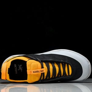 Puede incluir: Zapatilla deportiva negra y amarilla con puntera blanca. La zapatilla presenta cordones negros y el nombre de la marca "Arc'teryx" en la leng&uuml;eta y el lateral. La zapatilla est&aacute; sobre un fondo negro.