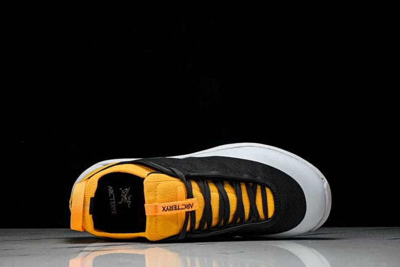 Puede incluir: Zapatilla deportiva negra y amarilla con puntera blanca. La zapatilla presenta cordones negros y el nombre de la marca "Arc'teryx" en la leng&uuml;eta y el lateral. La zapatilla est&aacute; sobre un fondo negro.