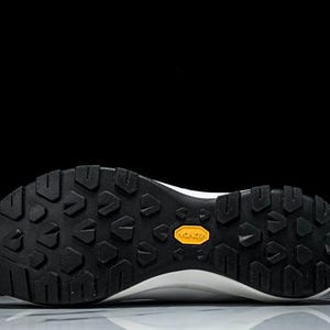 Puede incluir: Primer plano de la suela de una zapatilla deportiva blanca y negra. La suela es negra con un patr&oacute;n texturizado y un logotipo amarillo de Vibram. El calzado est&aacute; dise&ntilde;ado para actividades al aire libre.