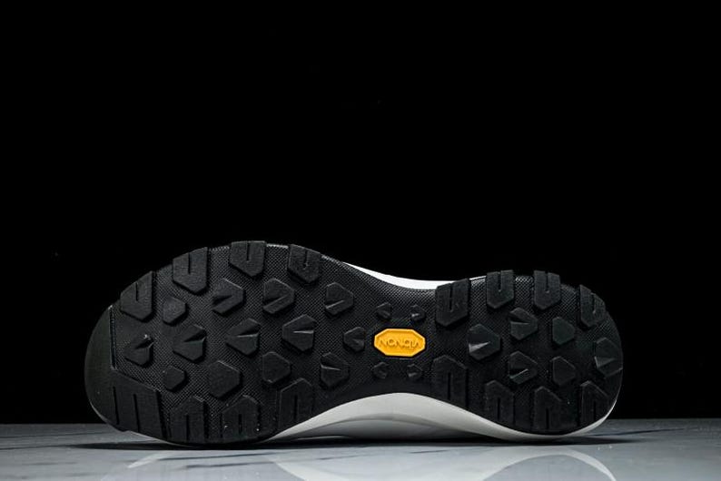 Puede incluir: Primer plano de la suela de una zapatilla deportiva blanca y negra. La suela es negra con un patr&oacute;n texturizado y un logotipo amarillo de Vibram. El calzado est&aacute; dise&ntilde;ado para actividades al aire libre.
