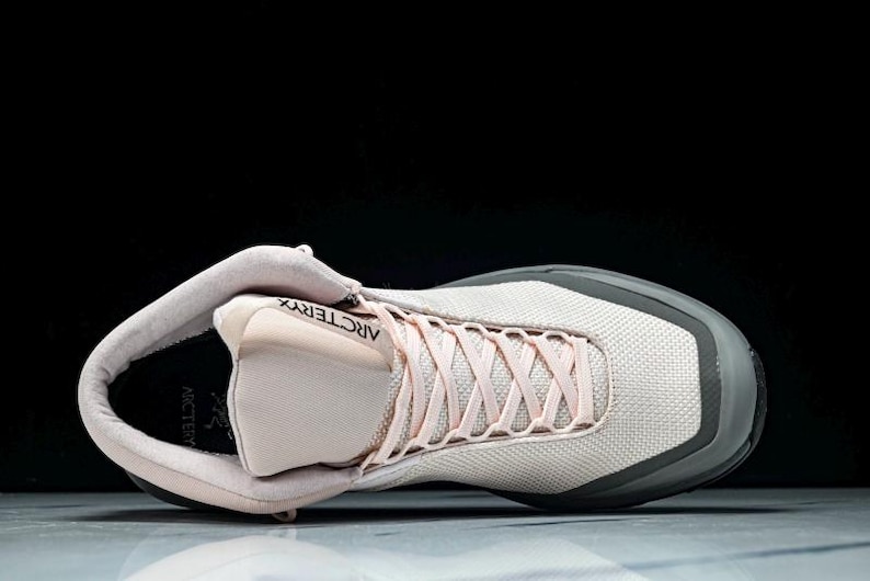 May include: A light pink and gray athletic shoe with the brand name "Arc'teryx" visible. The shoe has light pink laces and a gray sole. The shoe is designed for athletic activities.