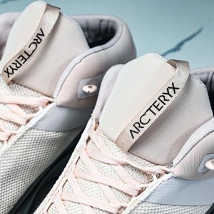 May include: Close-up of two light-colored athletic shoes. The shoes have a mesh-like texture on the sides and a light pink lace-up design. The tongue of each shoe has the word "ARCTERYX" printed in black.