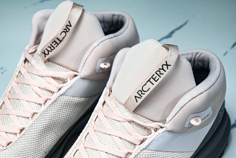 May include: Close-up of two light-colored athletic shoes. The shoes have a mesh-like texture on the sides and a light pink lace-up design. The tongue of each shoe has the word "ARCTERYX" printed in black.