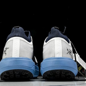 May include: A pair of athletic shoes, viewed from the back, featuring a navy blue heel and tongue, white mesh uppers, and light blue soles. The soles have a black tread pattern. A tag with white text hangs from one shoe. The shoes have a logo on the side.