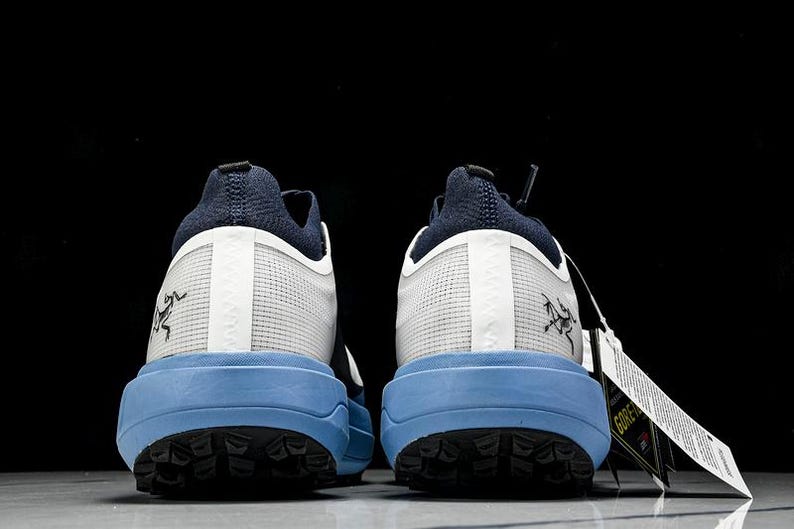 May include: A pair of athletic shoes, viewed from the back, featuring a navy blue heel and tongue, white mesh uppers, and light blue soles. The soles have a black tread pattern. A tag with white text hangs from one shoe. The shoes have a logo on the side.