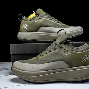May include: Two olive green athletic shoes with a thick sole are displayed. The shoes have a mesh and synthetic upper, with the word "SYLAN" printed on the side. The Arc'teryx logo is visible on the side of the shoe.