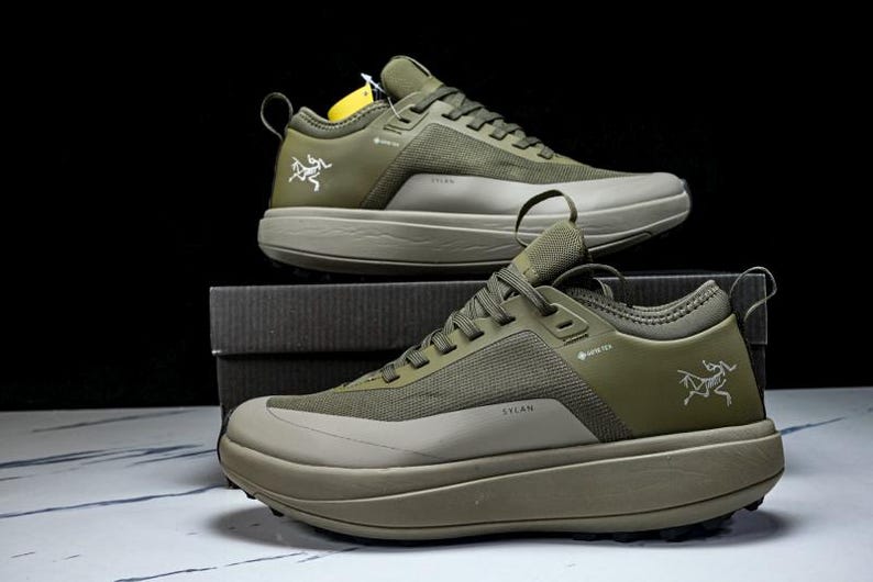 May include: Two olive green athletic shoes with a thick sole are displayed. The shoes have a mesh and synthetic upper, with the word "SYLAN" printed on the side. The Arc'teryx logo is visible on the side of the shoe.