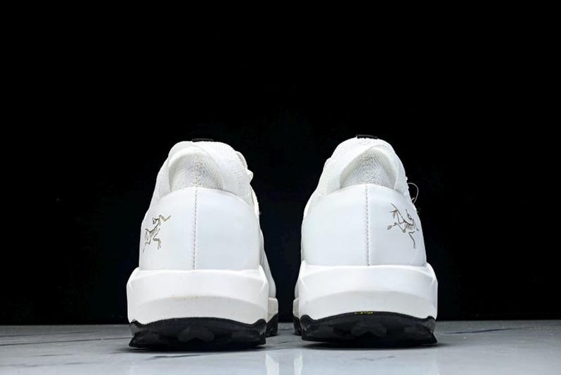 Puede incluir: Un par de zapatillas blancas con suelas negras, visto desde atr&aacute;s. Los zapatos tienen un dise&ntilde;o minimalista, talones texturizados y un peque&ntilde;o logotipo. Las suelas negras est&aacute;n texturizadas para el agarre.