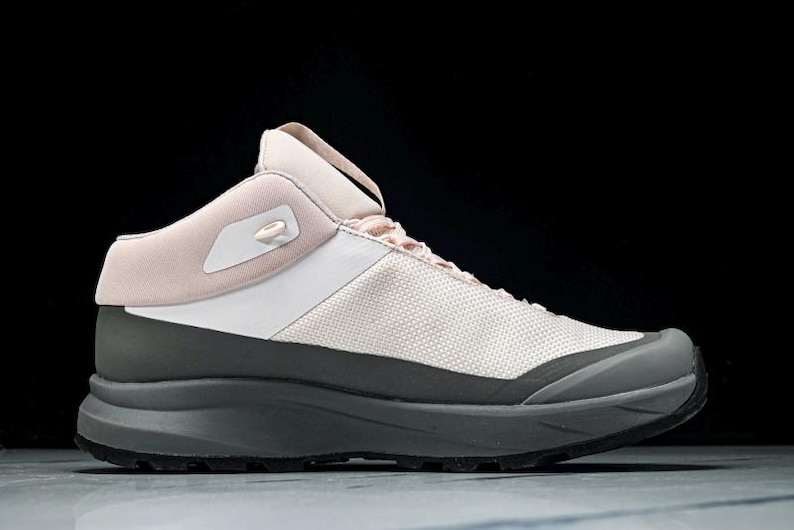 May include: A modern athletic shoe in a neutral color palette. The shoe features a light pink collar and tongue, a white midsection, and a gray base and sole. The design is sleek and contemporary, suitable for various activities.