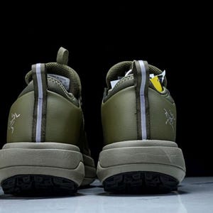 May include: A pair of olive green athletic shoes, viewed from the back. The shoes have a thick, tan sole and a textured tread. A reflective stripe runs down the back of each shoe. The brand logo is visible on the side.