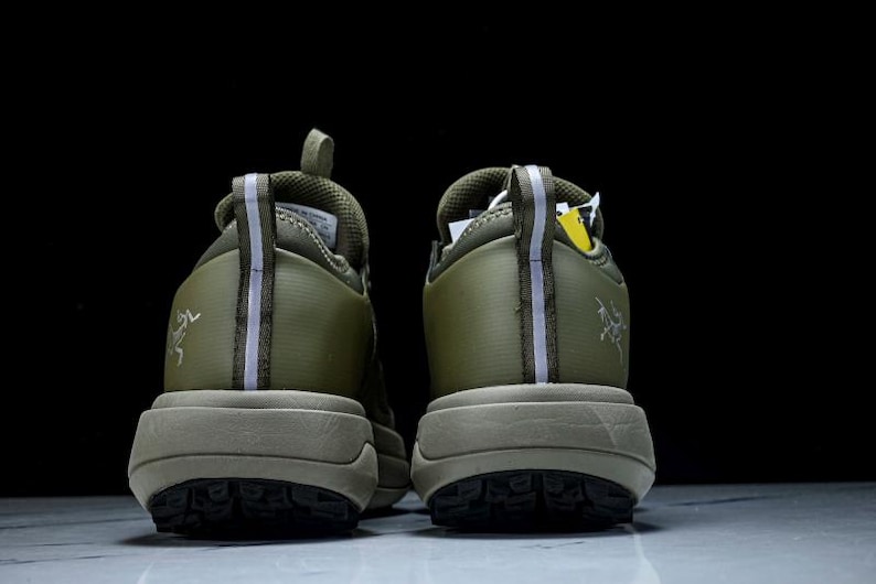 May include: A pair of olive green athletic shoes, viewed from the back. The shoes have a thick, tan sole and a textured tread. A reflective stripe runs down the back of each shoe. The brand logo is visible on the side.