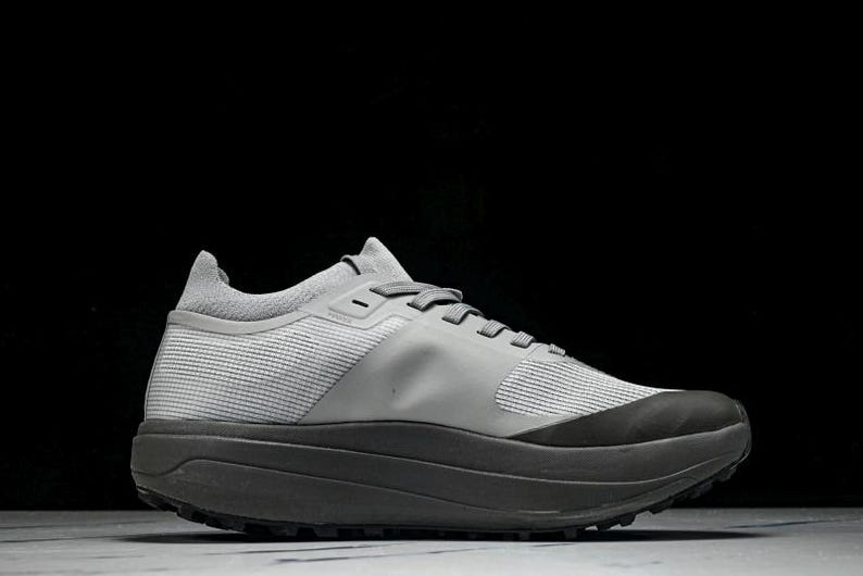 Puede incluir: Una zapatilla deportiva gris con suela gris oscuro y parte superior gris claro. La zapatilla tiene un dise&ntilde;o de malla texturizada en los laterales y un cuello acolchado. Los cordones son grises, sobre un fondo negro.