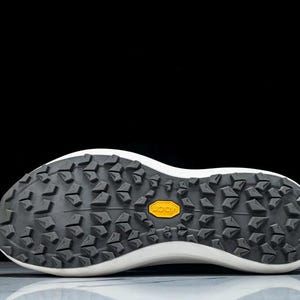 May include: Close-up of the sole of a white and black athletic shoe. The sole is black with a textured pattern and a yellow "Vibram" logo. The shoe's white midsole curves upwards.