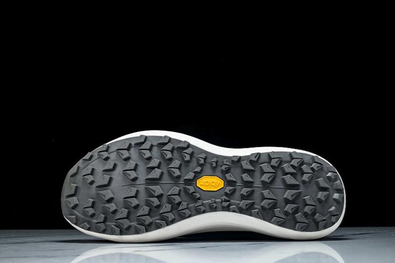 May include: Close-up of the sole of a white and black athletic shoe. The sole is black with a textured pattern and a yellow "Vibram" logo. The shoe's white midsole curves upwards.